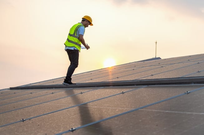 A worker in a yellow helmet inspects solar panels on a rooftop under a cloudy sky, with sunlight shining through.