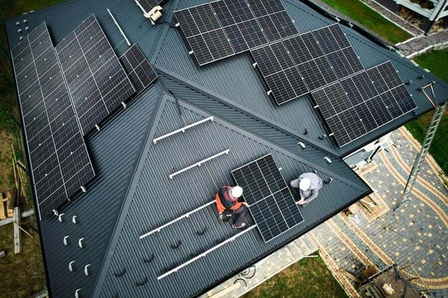 Aerial view of two workers installing solar panels on a dark metal house roof, with several panels already mounted and tools visible.