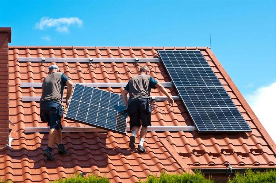 Two workers install solar panels on a red tiled roof under a clear blue sky.