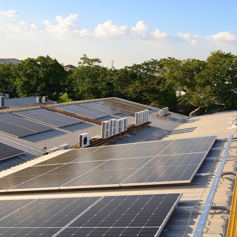Rows of solar panels installed on the rooftops of buildings, surrounded by trees under a partly cloudy sky.