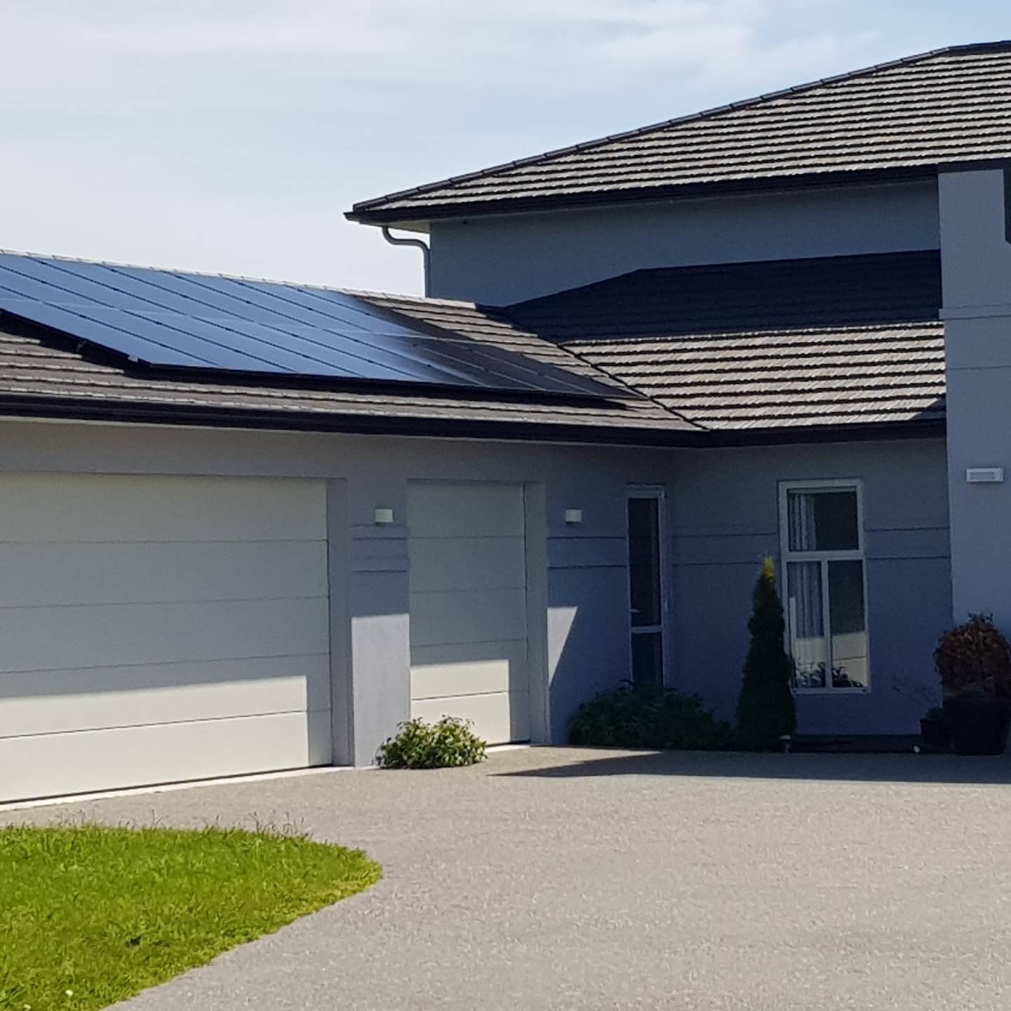 Modern two-story house with a double garage and solar panels installed on the garage roof, surrounded by a paved driveway and green lawn.